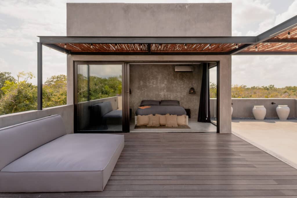 An outdoor terrace at Villa Niburu in Tulum features a lounging area with a large white sofa, and sliding glass doors open into a modern bedroom with minimalist decor.