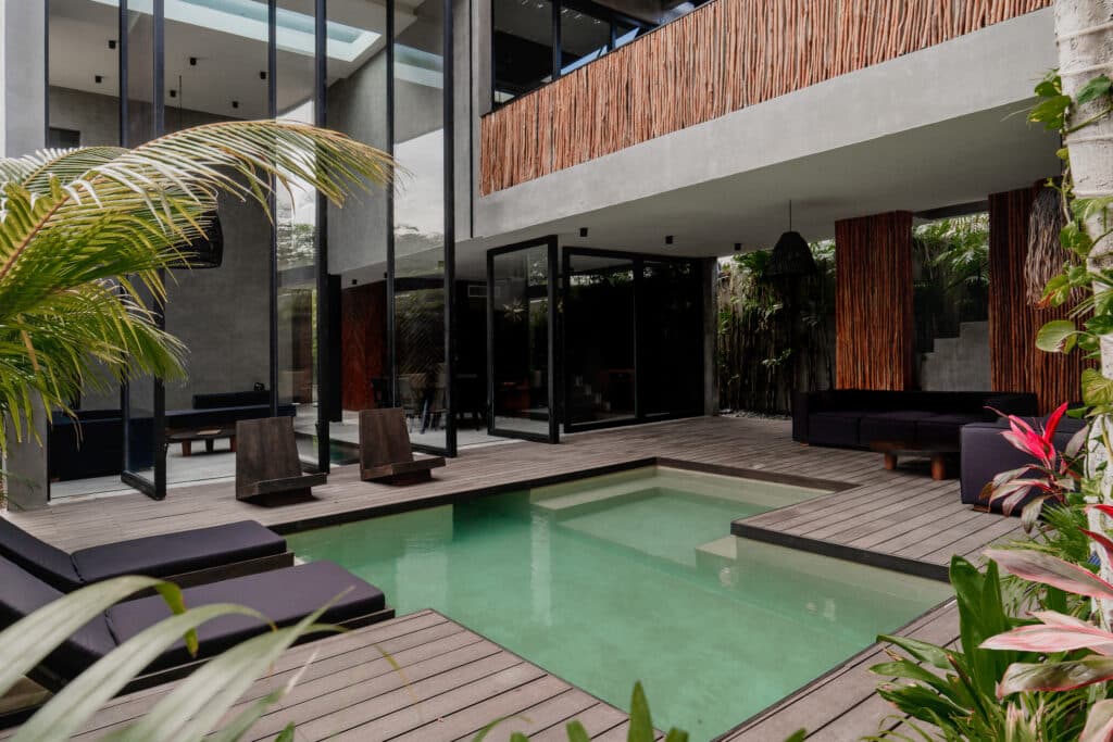 Modern Villa Niburu in Tulum features a sleek outdoor pool area with wooden decking, black lounge furniture, and glass doors opening to contemporary interior spaces.
