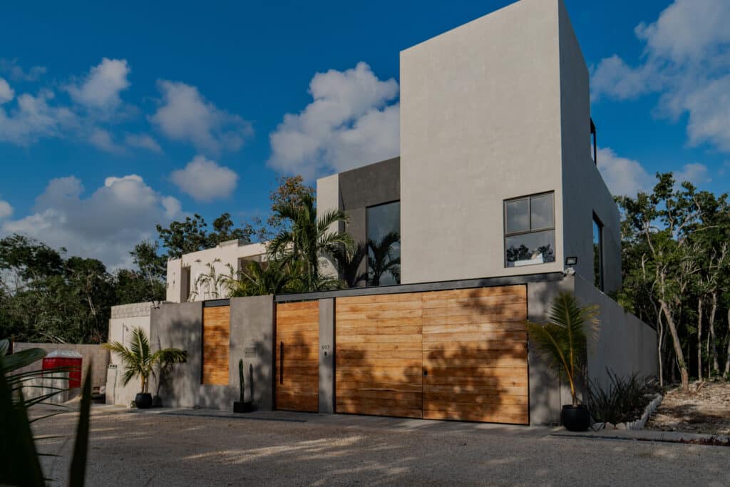 Modern white and gray villa with wooden gate, surrounded by tropical plants, located in Villa Niburu, Tulum's Region 15 under a partly cloudy sky.