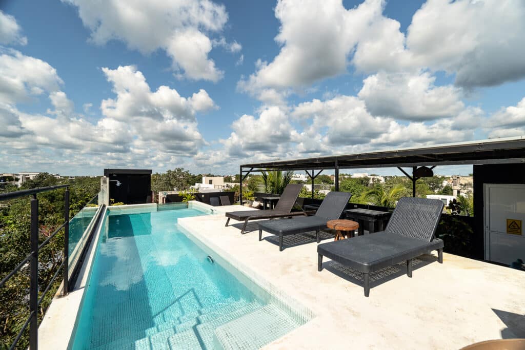 A rooftop pool area with lounge chairs and a shaded seating section overlooking trees and buildings in La Veleta, Tulum.