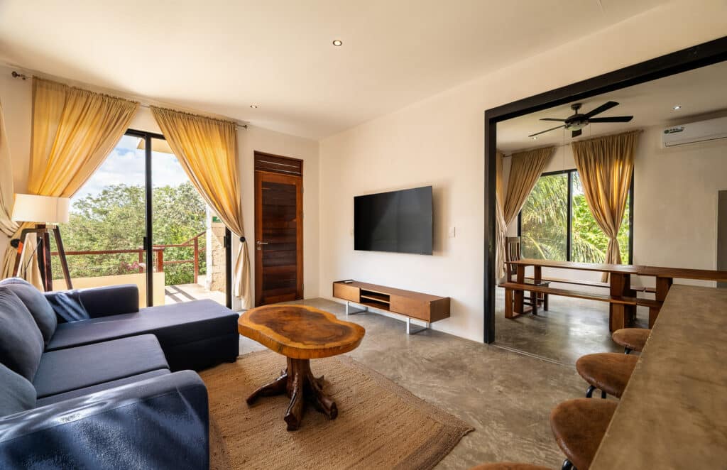 Interior view of the 10 Unit Hotel/Apartment Building in La Veleta, Tulum, showcasing a living area with a dark sofa, wooden coffee table, TV on the wall, and large windows with beige curtains.