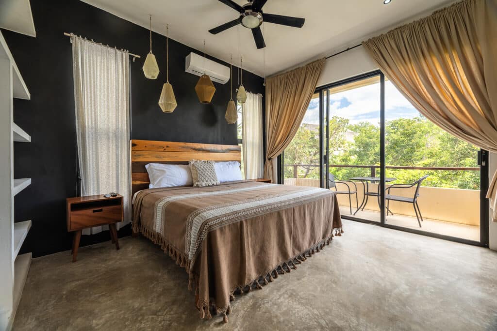 A bedroom in the 10 Unit Hotel/Apartment Building in La Veleta, Tulum, featuring a double bed, wooden accents, beige curtains, and a private balcony with outdoor seating.