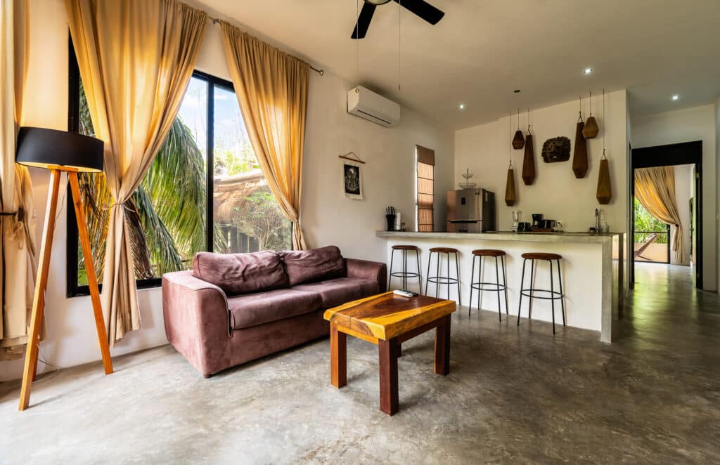 Living room with large windows, beige curtains, a mauve sofa, wooden coffee table, and a bar area with four stools in a contemporary Tulum apartment.