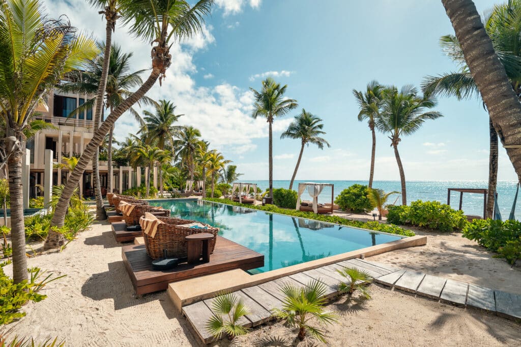 A view of a swimming pool surrounded by lounge chairs and palm trees at Bespoke D9 in Tankah Bay, Tulum, with the ocean and blue sky in the background.