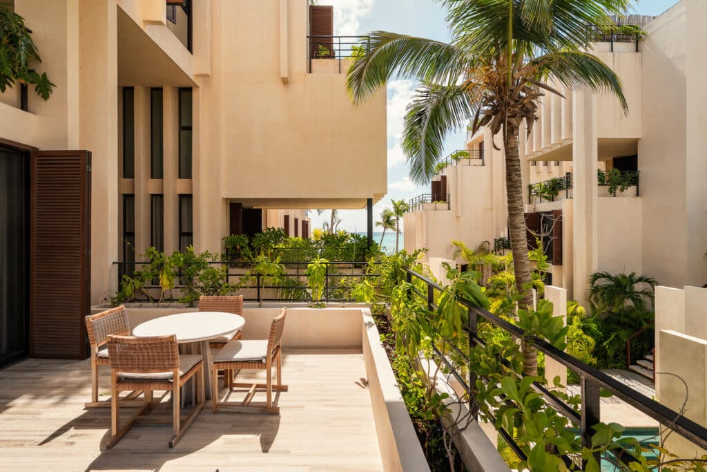 Balcony of a modern condo at Bespoke D9 in Tankah Bay, Tulum, featuring a small table, four chairs, lush greenery, a palm tree, and a view of neighboring buildings and the ocean.