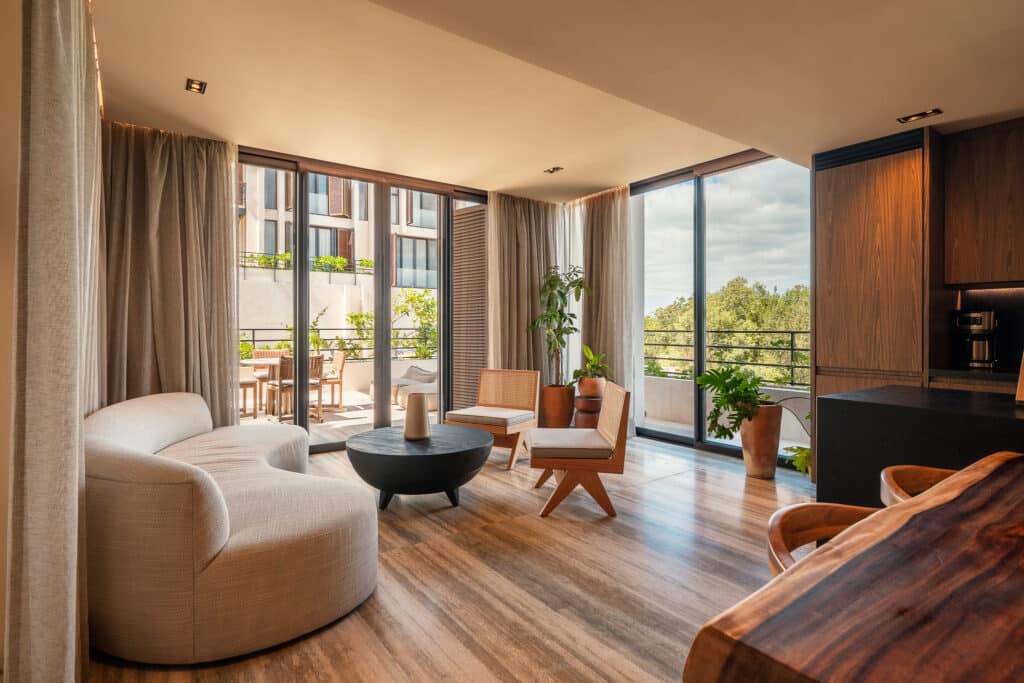 Modern living room with large glass doors opening to a balcony, featuring neutral furniture, wooden flooring, and potted plants at Bespoke D9 in Tulum's Tankah Bay.