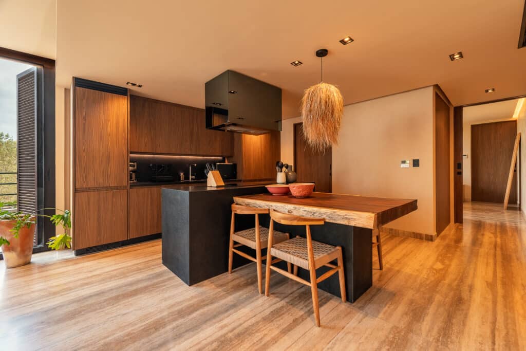 Interior view of a modern kitchen in the Bespoke D9 development in Tulum, featuring wooden cabinetry, a black kitchen island, and natural lighting from a nearby balcony.
