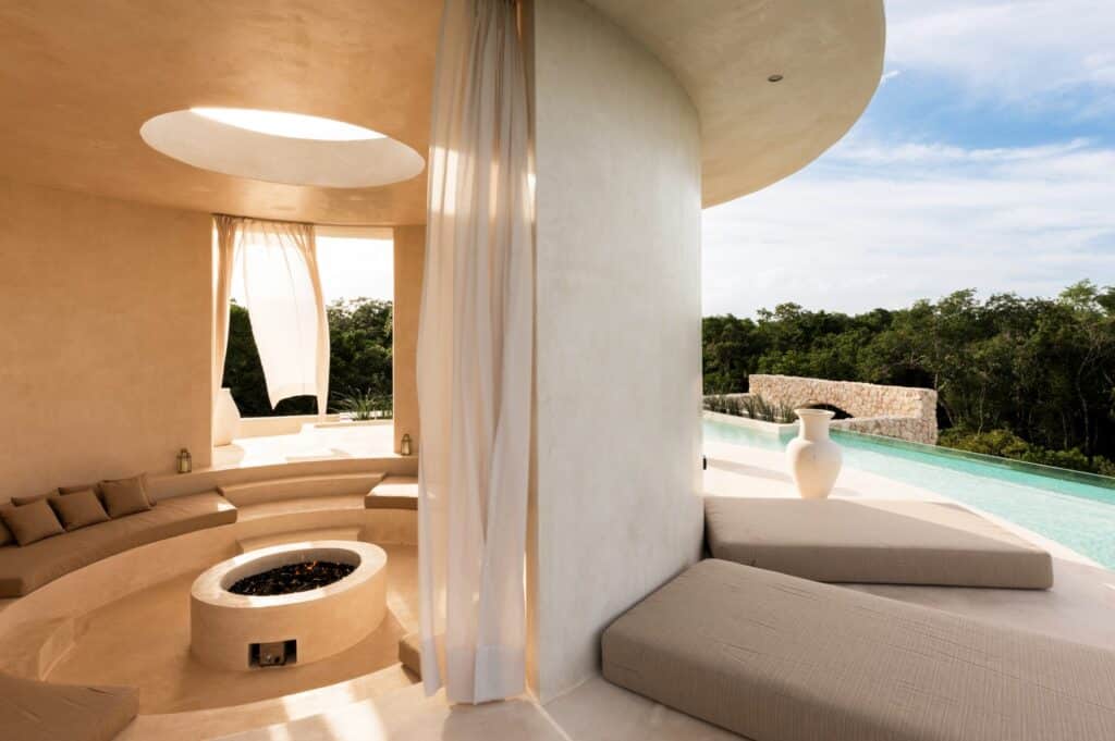 Interior view of Casa Arka in Tulum, featuring a cozy seating area around a fire pit with large windows, adjacent to an infinity pool overlooking lush greenery.