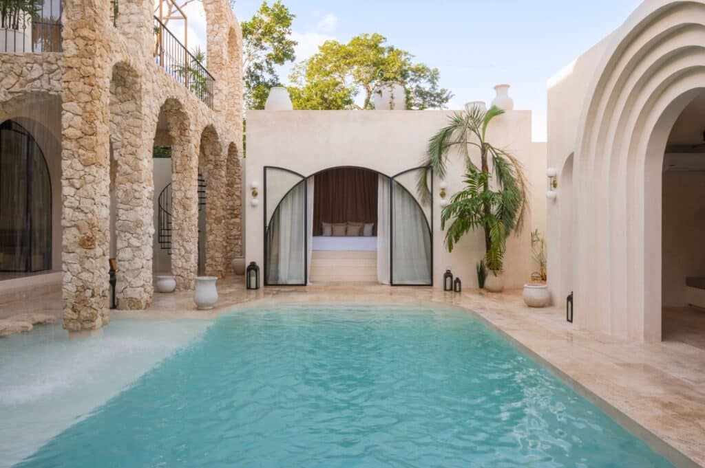 A luxurious house at Casa Arka in Tulum, featuring a pool with a shaded cabana area, surrounded by stone columns, lush plants, and modern arches.
