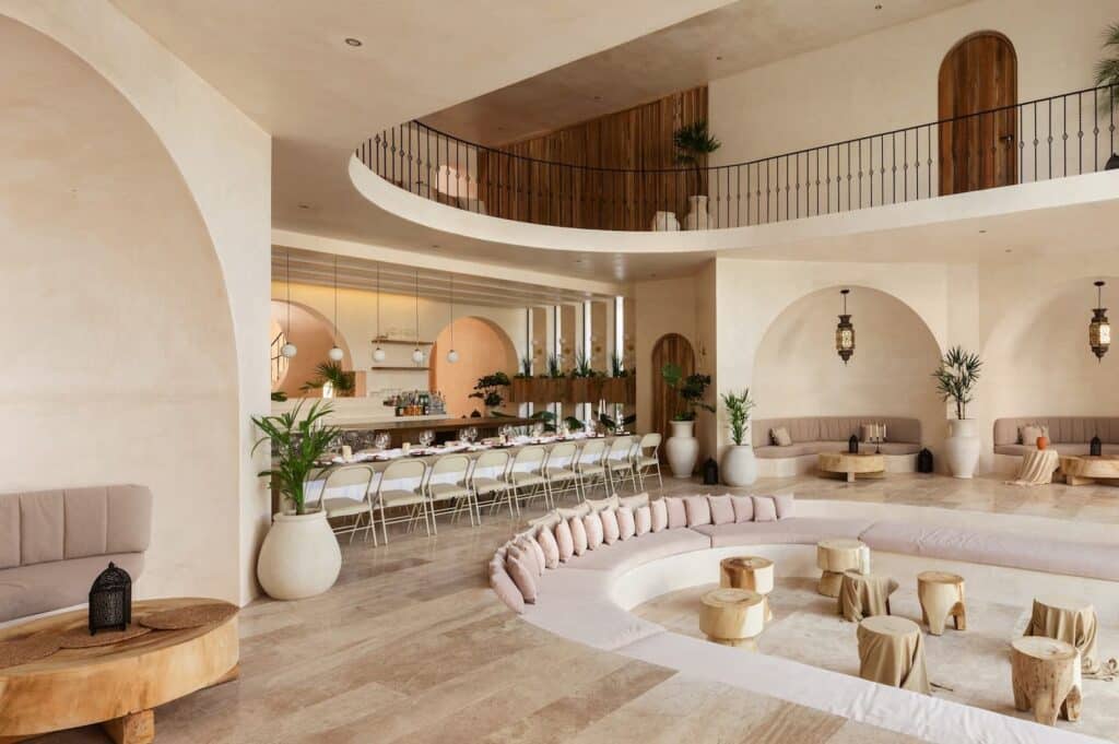 Interior view of Casa Arka in Tulum, featuring a spacious lounge area with curved seating, white walls, potted plants, decorative lighting, and a dining section with a long table.