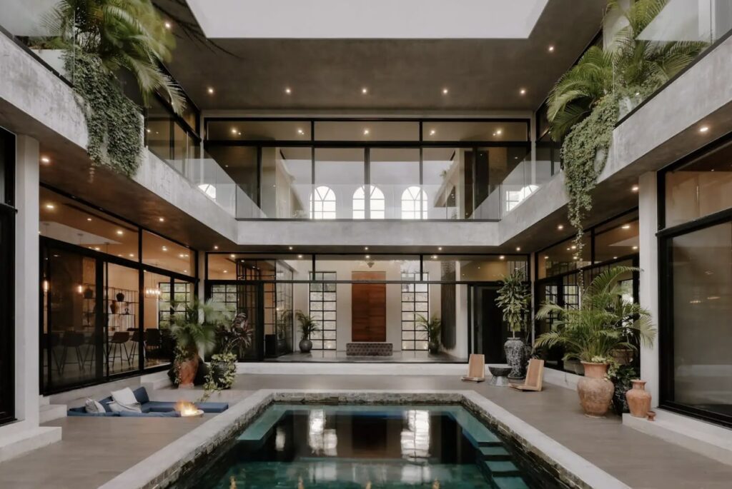 A spacious indoor courtyard at Casa Don Rey in Aldea Zama, Tulum, featuring a swimming pool, potted plants, large glass doors, and a double-height ceiling with modern decor.