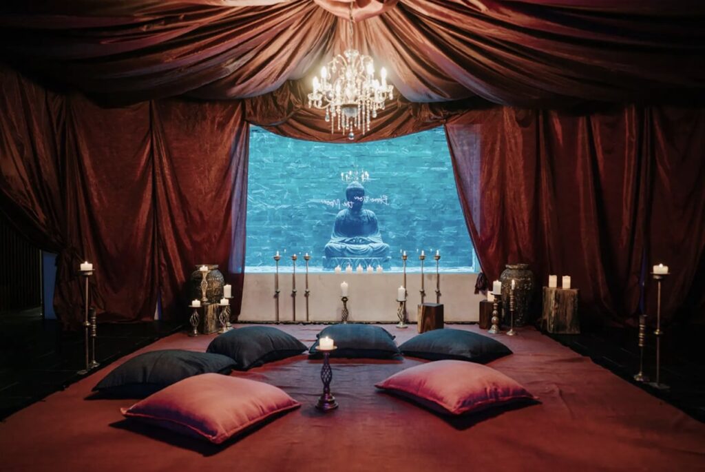 Interior view of Casa Don Rey in Aldea Zama, Tulum, featuring a cozy lounge area with floor cushions, candles, a chandelier, and a large Buddha statue visible through a window.