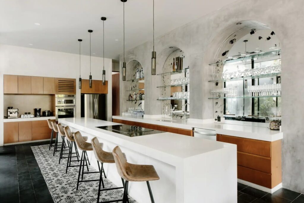 A modern kitchen in Casa Don Rey, Tulum, featuring a white island, wooden cabinetry, arched windows with glass shelves, and stylish pendant lighting.