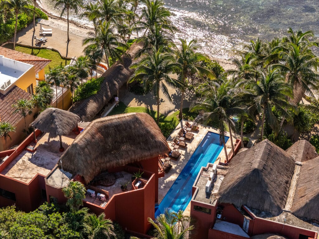 Aerial view of Jade Bay 8 beachfront development in Akumal, showing a swimming pool, thatched-roof structures, lush palm trees, and direct access to the sandy beach and turquoise water.
