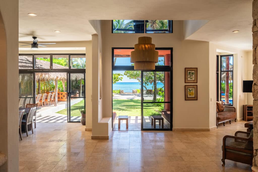 Interior view of Casa Zen Del Sol at Jade Bay 8 in Akumal, showcasing large windows opening to beachfront scenery with outdoor seating and lush greenery.