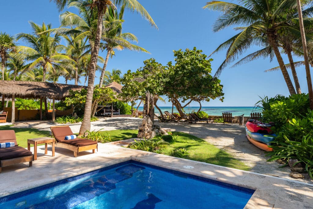 View of a beachfront property at Jade Bay 8 in Akumal featuring a pool, palm trees, lounge chairs, and kayaks with the ocean in the background on a sunny day.