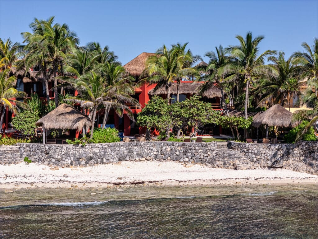 Beachfront Casa Zen Del Sol at Jade Bay 8 in Akumal, surrounded by palm trees and thatched-roof huts, with a stone wall separating the sandy beach from the property.