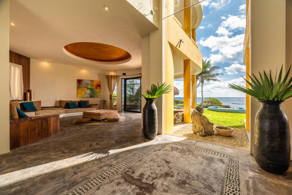 Interior view of Espirit del Mar Ocean Front House 2 in Punta Sur 5, Akumal, showing a spacious living area with large open doors opening to a beachfront view with palm trees and a clear sky.