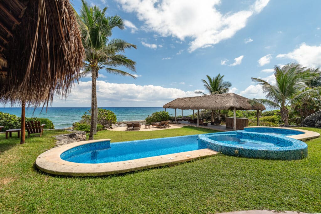 A view of a beachfront area at Punta Sur 5 in Akumal, featuring a small pool, palm trees, thatched-roof shaded lounge areas, and the ocean under a partly cloudy sky.