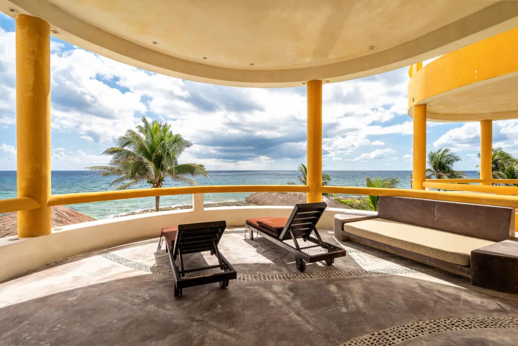 View from Espirit del Mar Ocean Front House 5 at Punta Sur 5 in Akumal, showcasing a curved balcony with lounge chairs and a view of the ocean and palm trees under a partly cloudy sky.
