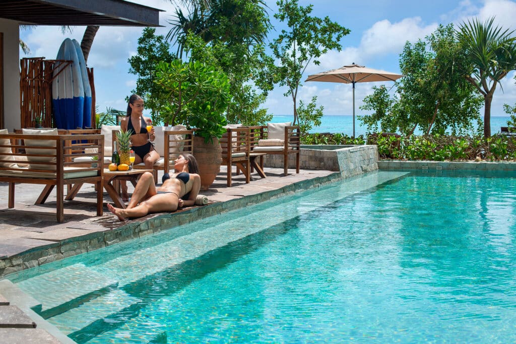 Two women relax by a pool at Tankah Bay 21 Room Beachfront Hotel in Tulum, surrounded by lush greenery with the ocean visible in the background.