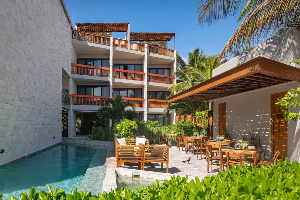 A view of the Tankah Bay 21 Room Beachfront Hotel in Tulum showing a swimming pool, outdoor dining area, lush greenery, and a modern building with multiple balconies under a clear blue sky.