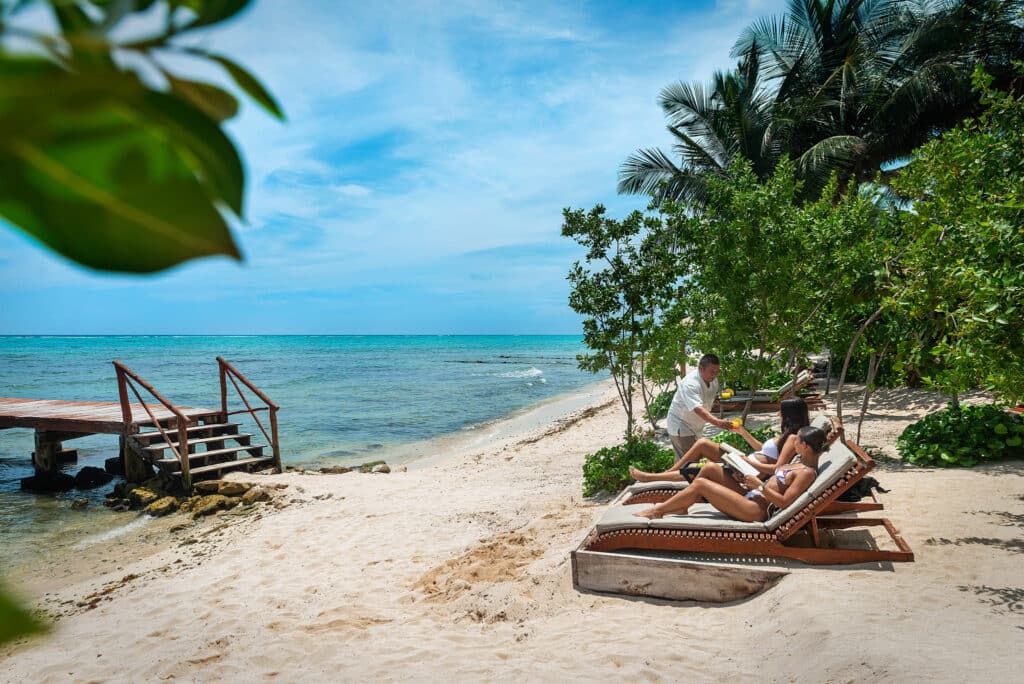 A beachfront scene at Tankah Bay in Tulum showing two women lounging on deck chairs under trees with clear blue sky and calm ocean in the background.