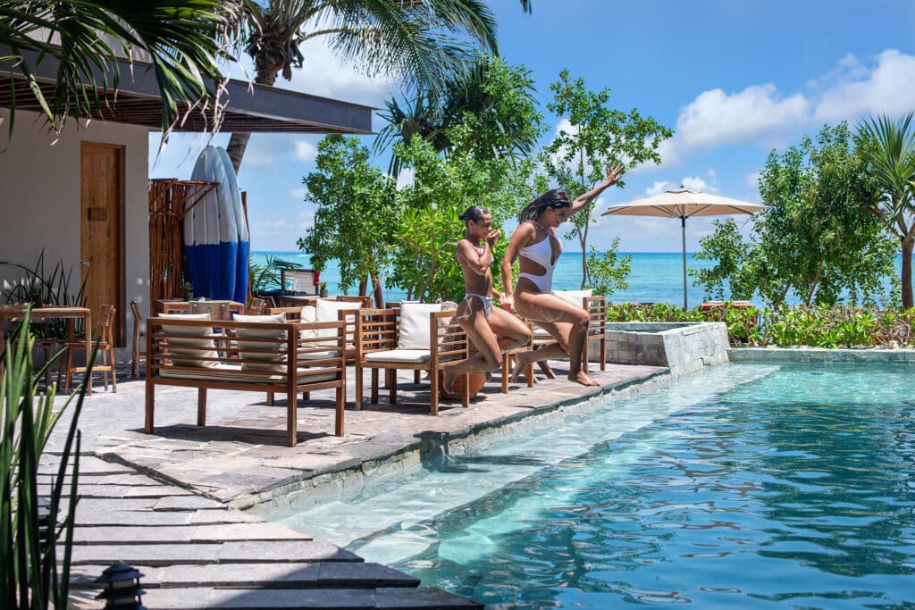 Two women in swimsuits jump into a pool at Tankah Bay 21 Room Beachfront Hotel in Tulum, surrounded by greenery, with the ocean visible in the background under a blue sky.