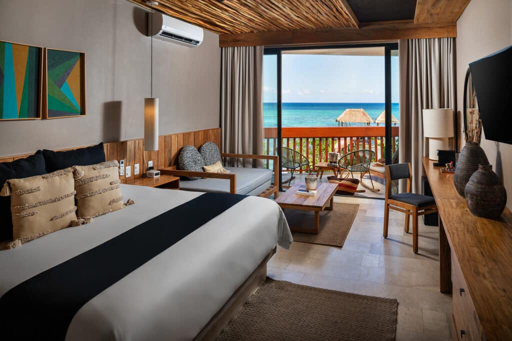 Interior view of a beachfront hotel room at Tankah Bay in Tulum, featuring a bed, sitting area, wooden furniture, and a balcony with ocean views.