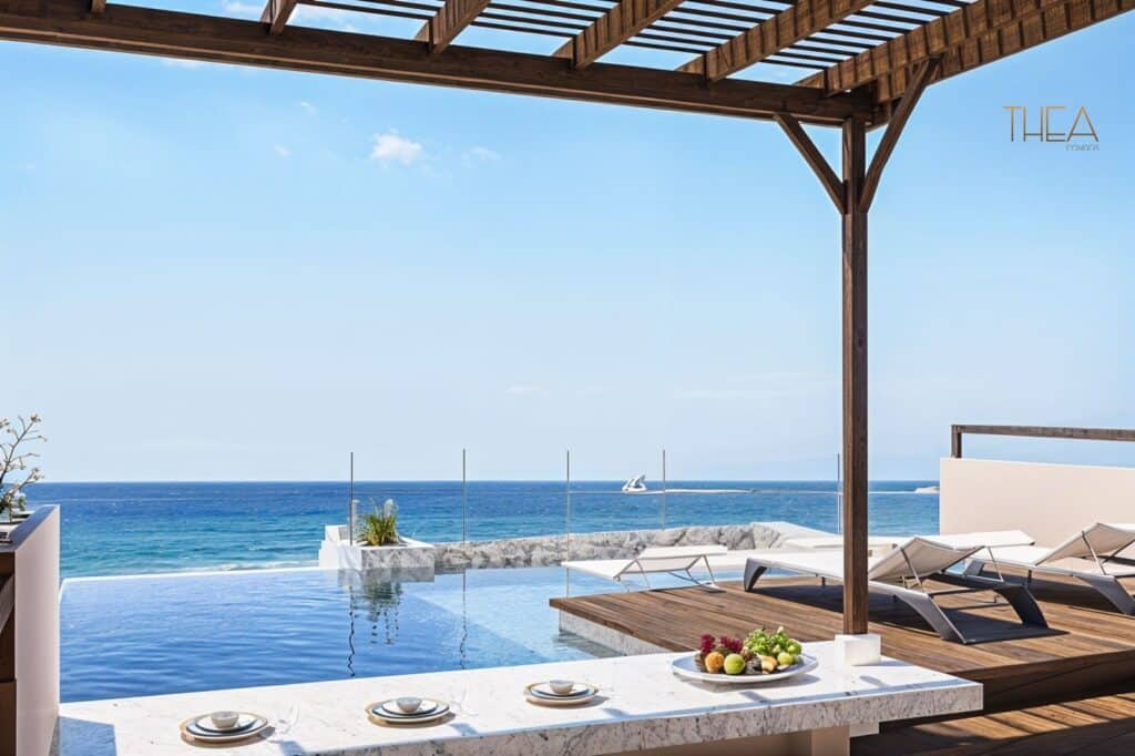 A view from Thea 502 in Puerto Morelos shows an infinity pool overlooking the ocean with lounge chairs and a wooden pergola, set against a clear blue sky and calm sea.
