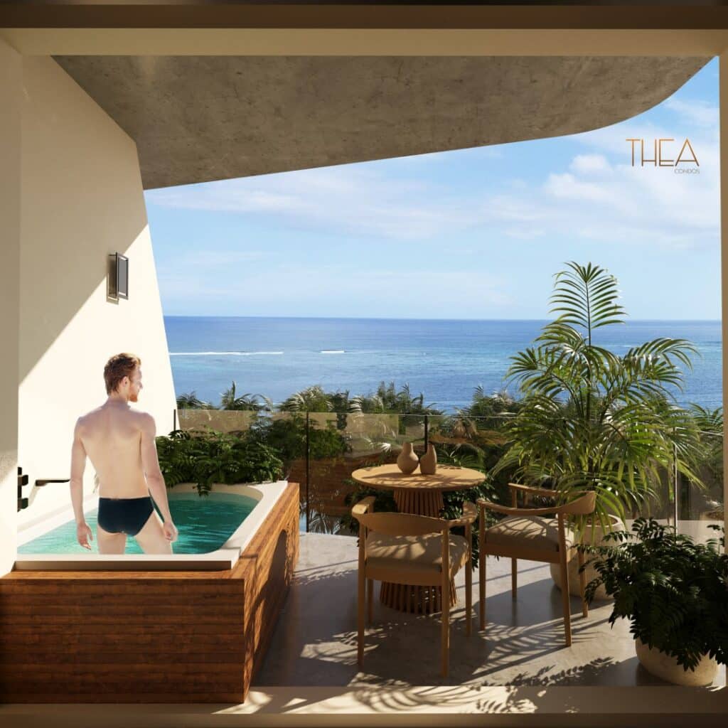 A man relaxes in a hot tub on a balcony overlooking the ocean with lush greenery and a clear blue sky at Thea 502 in Puerto Morelos.