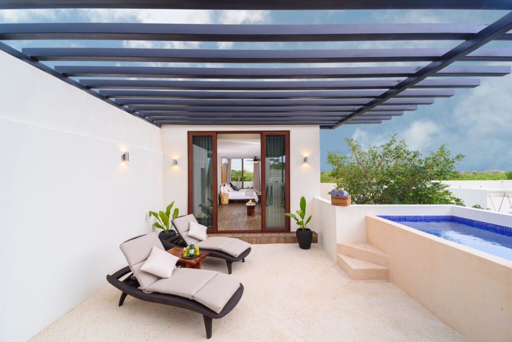 A rooftop terrace at the 25 Room Boutique Hotel in Aldea Zama, Tulum, featuring lounge chairs, potted plants, a small pool, and a view into a bedroom through glass doors.