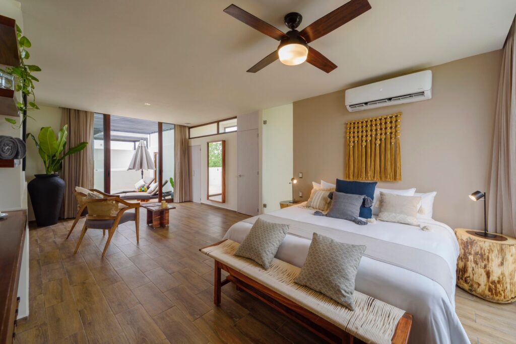 A spacious hotel room at the 25 Room Boutique Hotel in Aldea Zama, Tulum, featuring a large bed, wooden furniture, a ceiling fan, and an indoor-outdoor sitting area.