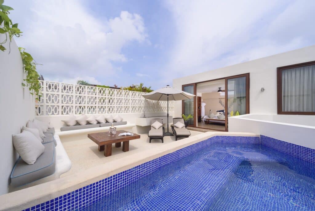 A spacious outdoor patio at the 25 Room Boutique Hotel in Aldea Zama, Tulum, featuring a small pool, lounge chairs, an umbrella, and seating area with pillows, with a view into a bedroom.