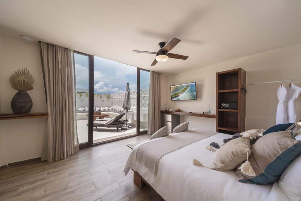 A bedroom in the 25 Room Boutique Hotel in Aldea Zama, Tulum, featuring a large bed with pillows, a ceiling fan, a wall-mounted TV, and a sliding door leading to an outdoor lounge area with sunbeds.