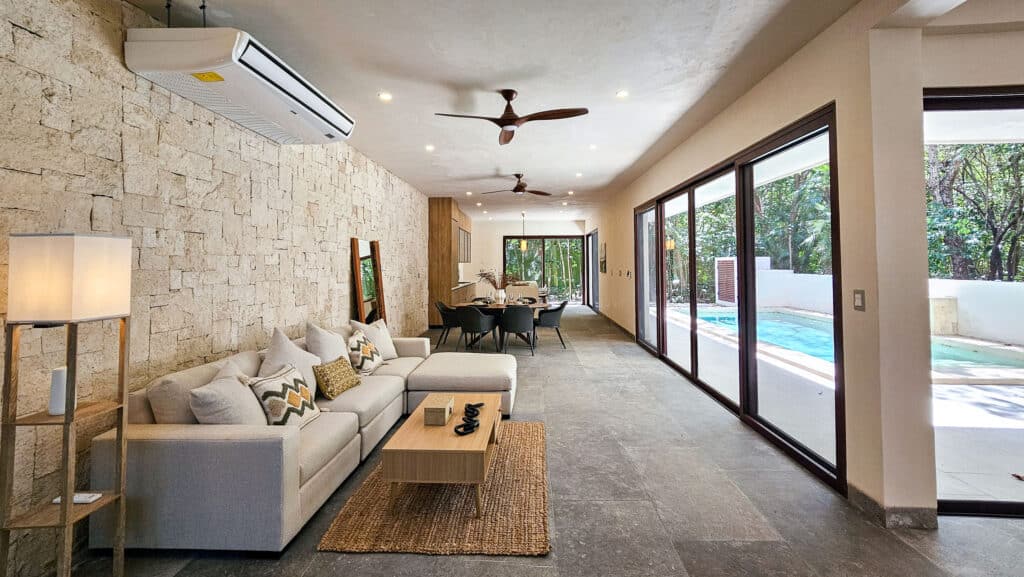 Living room and dining area of Villa Coral in Aldea Zama, Tulum, featuring a stone accent wall, large sliding glass doors leading to a pool area, and modern decor.