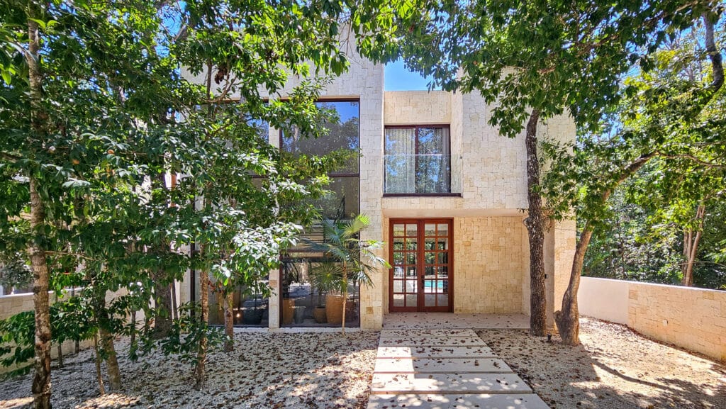 Front view of Villa Coral in Aldea Zama, Tulum, featuring a modern stone façade, large glass windows, wooden door, and lush trees surrounding the entrance.