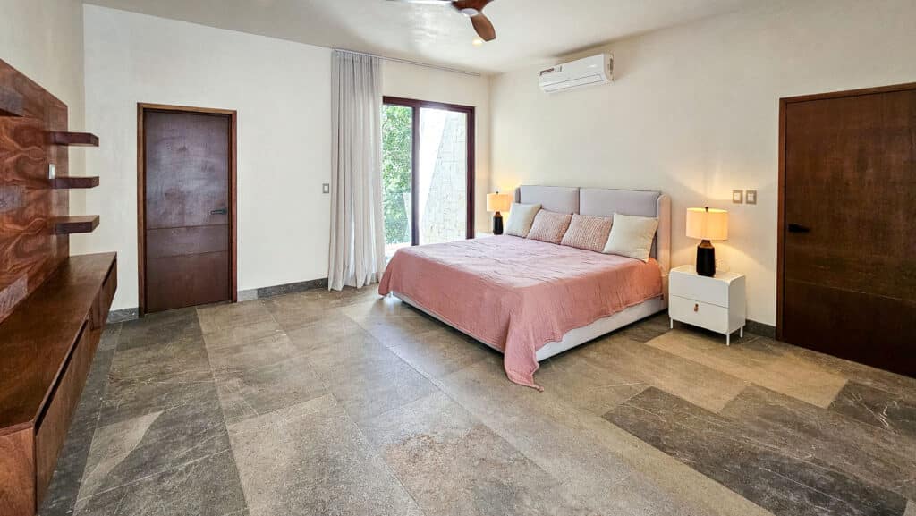 A bedroom in Villa Coral, Aldea Zama, Tulum, featuring a large bed with beige and blush bedding, wooden accents, a balcony view of greenery, and modern lighting fixtures.