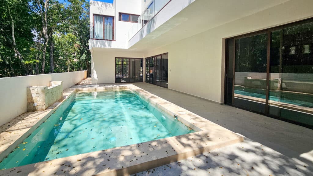 A modern white villa with a private pool overlooks lush trees in Aldea Zama, Tulum, within Villa Coral development.