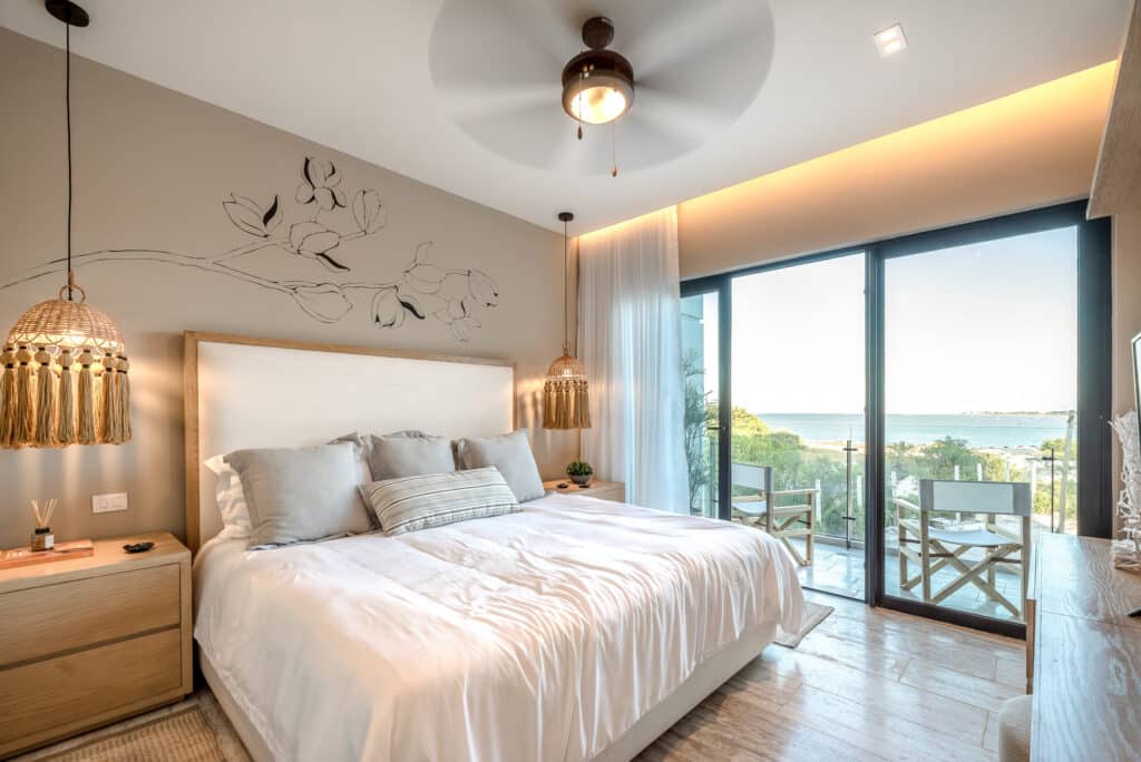 A bedroom at Bonanza PH in Progreso Marina Front features a cozy bed with neutral linens, wooden accents, decorative hanging lamps, and large glass doors opening to a balcony with ocean views.