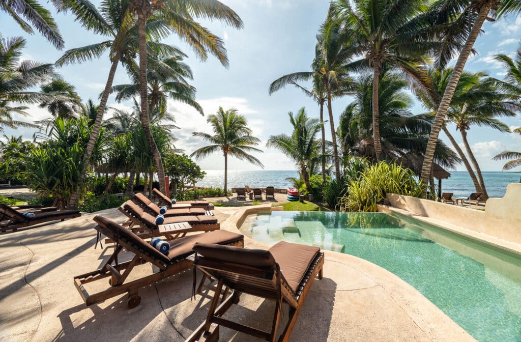 View of Jade Bay 9 beachfront development in Akumal featuring a pool with lounge chairs, palm trees, and ocean in the background under a partly cloudy sky.