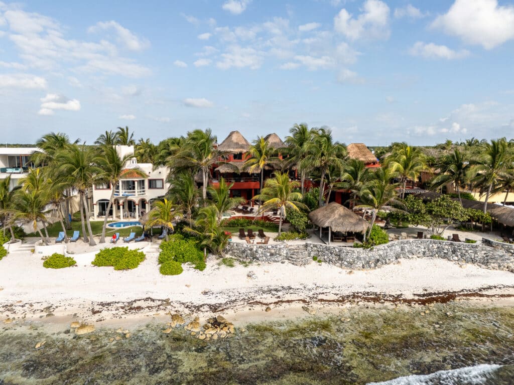 A beachfront development in Akumal featuring lush palm trees, white sandy beaches, thatched-roof structures, and modern villas with ocean views at Jade Bay 9.