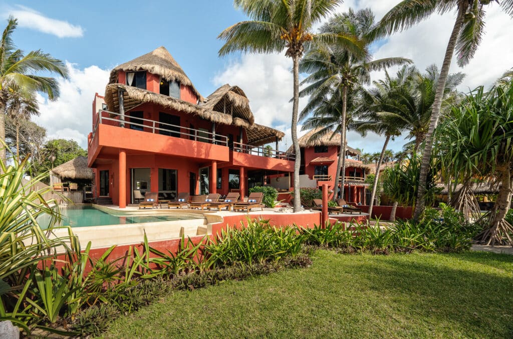 A beachfront development in Akumal featuring a coral-colored building with thatched roofs, surrounded by palm trees and lush greenery, with a pool and lounge chairs outside.