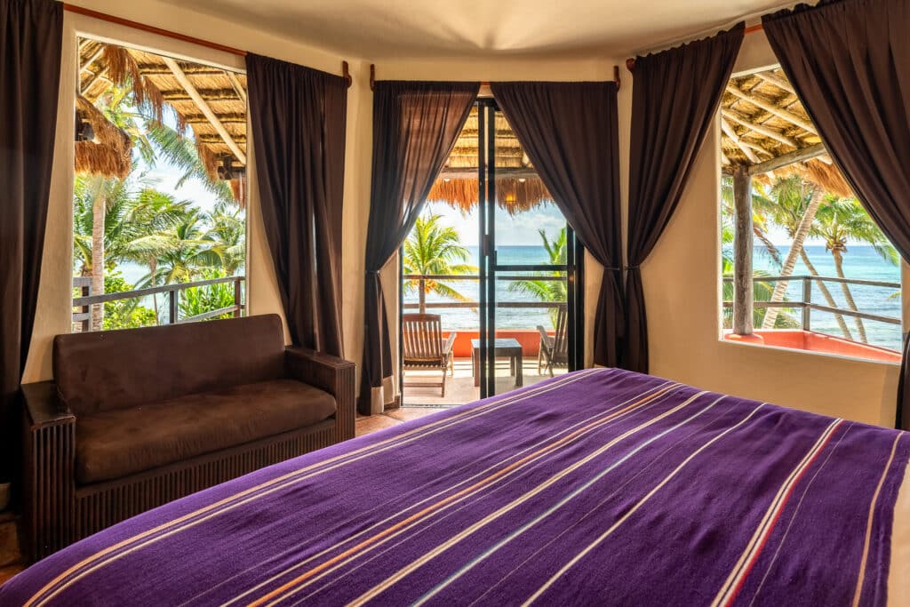 Interior view of a beachfront room at Jade Bay 9 in Akumal, showing a bed with a purple blanket, a brown sofa, dark curtains, and a balcony with ocean and palm tree views.