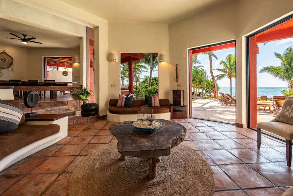 Interior view of Jade Bay 9, a beachfront property in Akumal, showing a living room with terracotta tile flooring, tropical decor, and open doors leading to a beachside patio with loungers and palm trees.