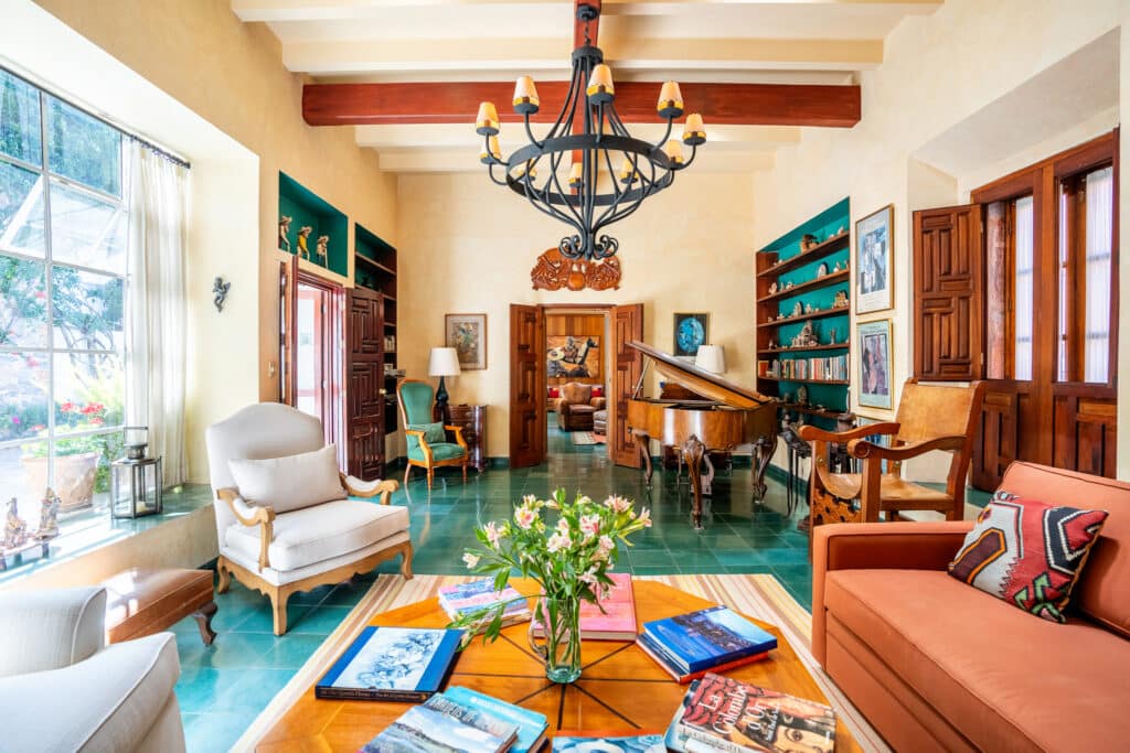 Living room in Casa Carolina, San Miguel de Allende, with a grand chandelier, wooden beams, a grand piano, built-in shelves, and colorful furniture.