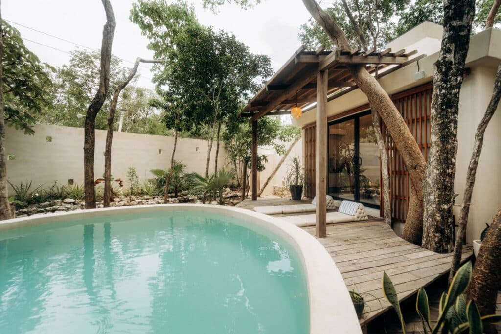 A cozy house at Casa Oox in Tulum featuring a small pool, surrounded by trees and greenery, with a shaded wooden deck and sliding glass doors.