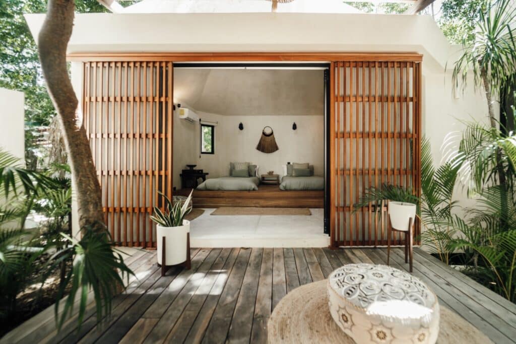 View of Casa Oox in Tulum's Region 15 showing an open-air bedroom with twin beds, surrounded by wooden slats, plants, and natural light.