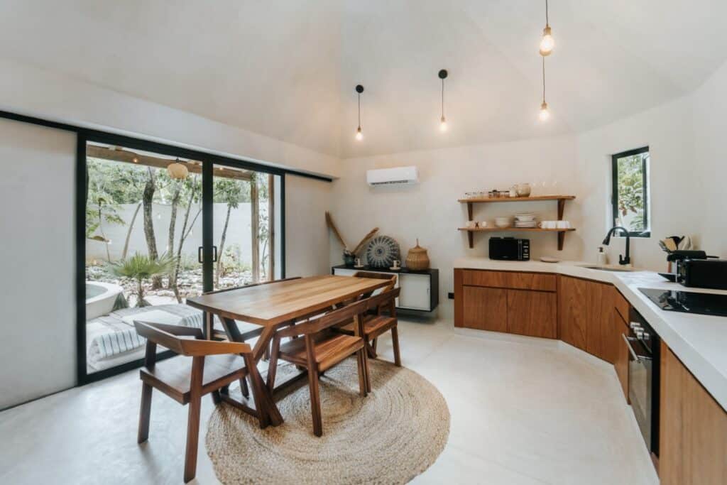 Interior of Casa Oox in Tulum, featuring a modern kitchen with wooden cabinets, open shelving, a dining area with a wooden table, and large sliding glass doors opening to a garden.
