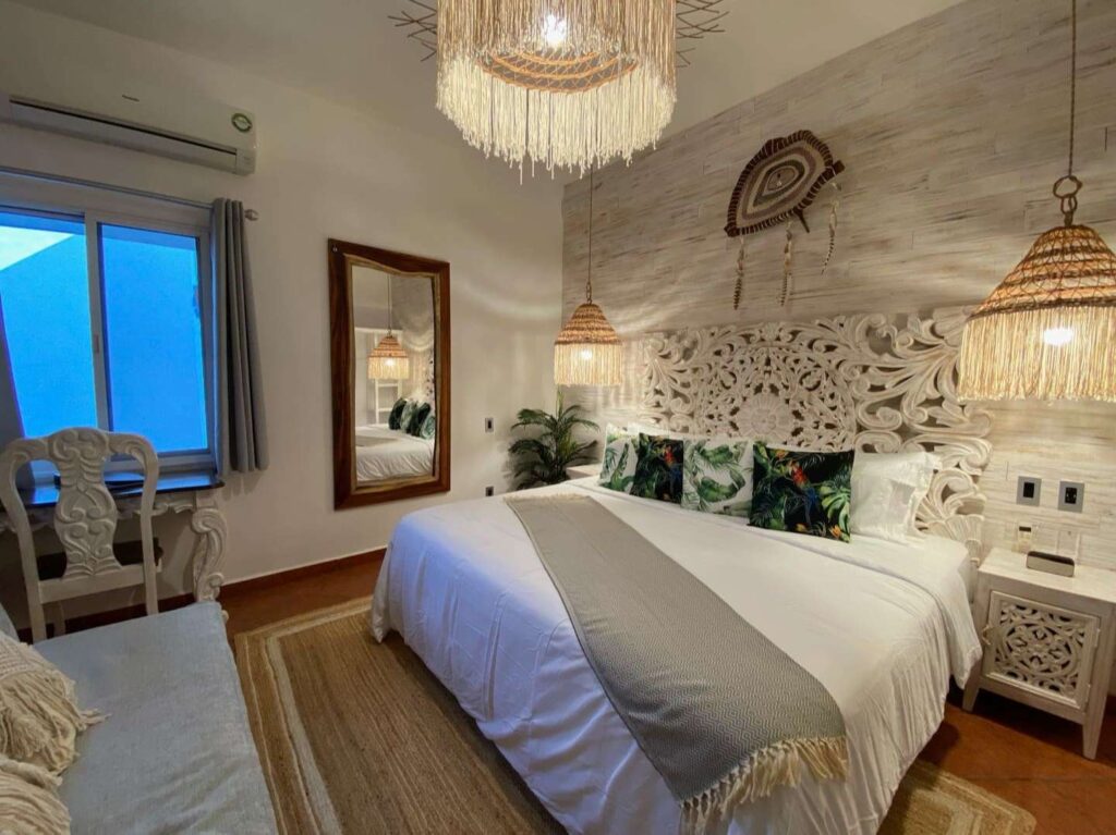 A beautifully decorated bedroom in Castillo 1 at Centro Tulum 6 Room Hotel in Tulum, showcasing a carved white headboard, tropical pillows, and woven lighting fixtures.